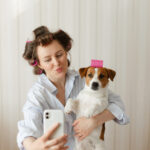 Beautiful young woman wearing pajamas with pink hair curlers holds a Jack Russell Terrier dog and takes a selfie on a smartphone at home on a sunny morning. Girl with her cute puppy, both in curlers, takes a photo on their phone